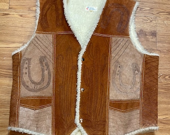May include: A brown suede vest with a faux fur lining. The vest has a patchwork design with horseshoes and a woodgrain pattern. The vest has a button closure and two pockets.