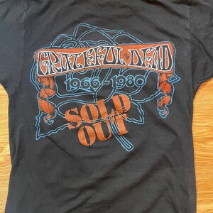 Puede incluir: Camiseta negra con un gr&aacute;fico azul y marr&oacute;n de una rosa con el texto "Grateful Dead 1966-1980 Sold Out" en ella.