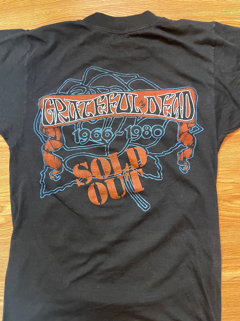 Puede incluir: Camiseta negra con un gr&aacute;fico azul y marr&oacute;n de una rosa con el texto "Grateful Dead 1966-1980 Sold Out" en ella.