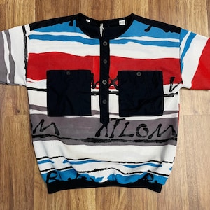 Puede incluir: Camiseta de manga corta con un diseño colorido. La camisa presenta un cuello y tapeta de botones negros, dos bolsillos cuadrados negros y un patrón de rayas rojas, azules, blancas y grises. La palabra "NATION" está impresa en la parte delantera.