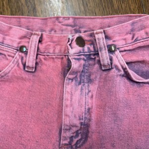 May include: Close-up of pink denim jeans with a faded, distressed look. The jeans feature a button closure, belt loops, and front pockets. The fabric has a mottled appearance with darker purple accents.