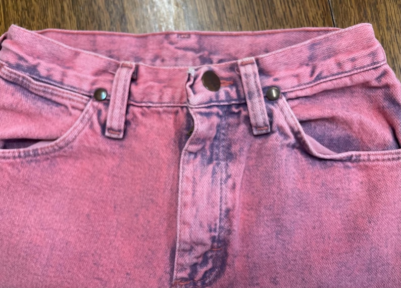 May include: Close-up of pink denim jeans with a faded, distressed look. The jeans feature a button closure, belt loops, and front pockets. The fabric has a mottled appearance with darker purple accents.