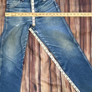 May include: Blue denim jeans with a classic five-pocket design. The waist measures approximately 15 inches across, and the inseam is about 28 inches. The jeans are laid flat on a wooden surface, showcasing their vintage style.