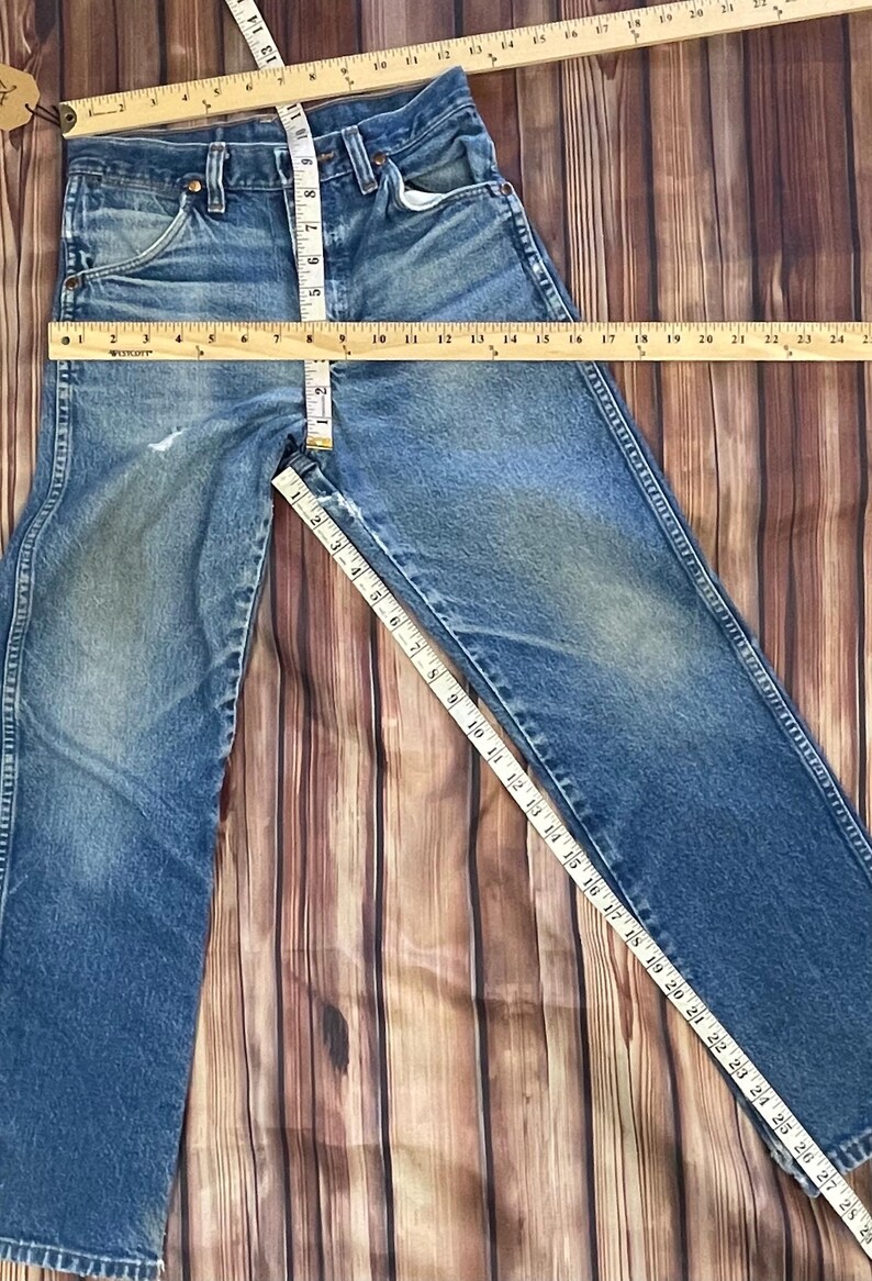 May include: Blue denim jeans with a classic five-pocket design. The waist measures approximately 15 inches across, and the inseam is about 28 inches. The jeans are laid flat on a wooden surface, showcasing their vintage style.