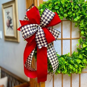May include: A vibrant green wreath adorned with a large red bow featuring a black and white checkered pattern. The wreath is displayed on a light-colored wooden screen, creating a striking contrast. The bow's long red streamers add a touch of elegance.