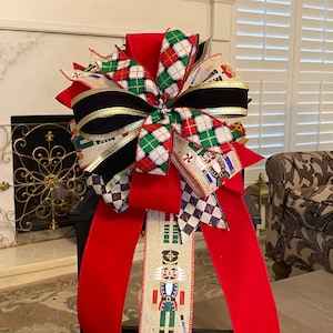 Puede incluir: Un lazo de terciopelo rojo con cinta de cuadros negros y blancos, una cinta de cascanueces y una cinta de terciopelo negro con ribete dorado. El lazo está atado alrededor de una linterna negra.