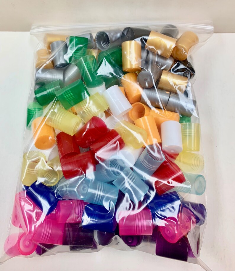 May include: A clear plastic bag filled with a variety of colorful plastic bottle caps. The caps are in various shapes and sizes, including round, square, and rectangular. The colors include red, orange, yellow, green, blue, purple, and white.