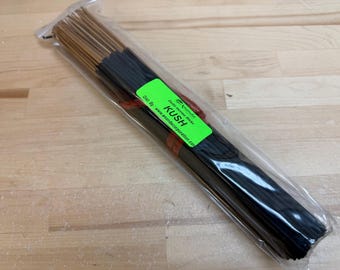 May include: A bundle of incense sticks in a clear plastic wrapper. The sticks are dark brown, with a green label that reads "KUSH". The packaging is on a light wood surface.