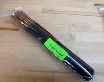 May include: A clear plastic package containing incense sticks. The sticks are black with gold tips and a green label that reads "Peppermint". The package is on a light-colored wooden surface.