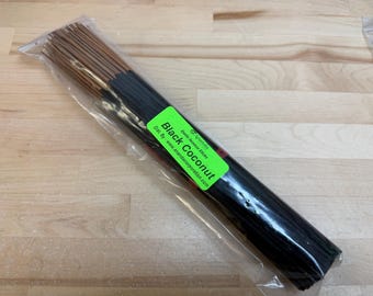May include: A bundle of black incense sticks in a clear plastic bag. A green label reads "Black Coconut" with additional text. The incense sticks are dark brown at the top and black at the bottom. The packaging is on a light wood surface.