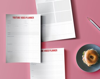 May include: Three white sheets of paper with the text "YOUTUBE VIDEO PLANNER" and "UBE VIDEO PLANNER" printed in red. The sheets are on a pink surface with a gray plate and a pencil.