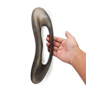 Puede incluir: Una escultura abstracta de color bronce, con un diseño liso y curvo y una gran abertura central. La forma alargada es sostenida por una mano sobre un fondo blanco, mostrando su forma única y acabado metálico.
