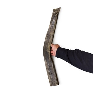 May include: A long, curved piece of wood with a dark brown and light brown color. The wood has a rough, textured surface. A hand is holding the wood.