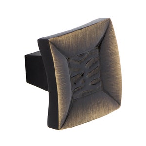 May include: A square cabinet knob with a brushed antique brass finish. The knob has a raised, textured centre and a black base.