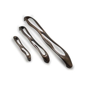 Può includere: Tre maniglie per mobili color bronzo di varie dimensioni. Ogni maniglia ha un design allungato e aperto con un manico curvo e una finitura metallica liscia. Le maniglie sono disposte diagonalmente su uno sfondo bianco.