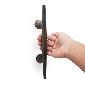 May include: A dark bronze door pull with a tapered design. The handle is held against a white background, showcasing its sleek, modern aesthetic. The pull is attached to two circular mounting plates.
