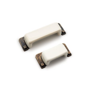 May include: Two white ceramic cabinet pulls with antique brass accents. The pulls are rectangular in shape with a slight curve on the top and bottom. The smaller pull is on top of the larger pull.