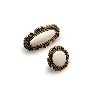 Peut inclure: Deux boutons de cabinet ornés de couleur or avec des inserts en céramique blanche. Le plus grand bouton est de forme ovale, tandis que le plus petit bouton est rond.