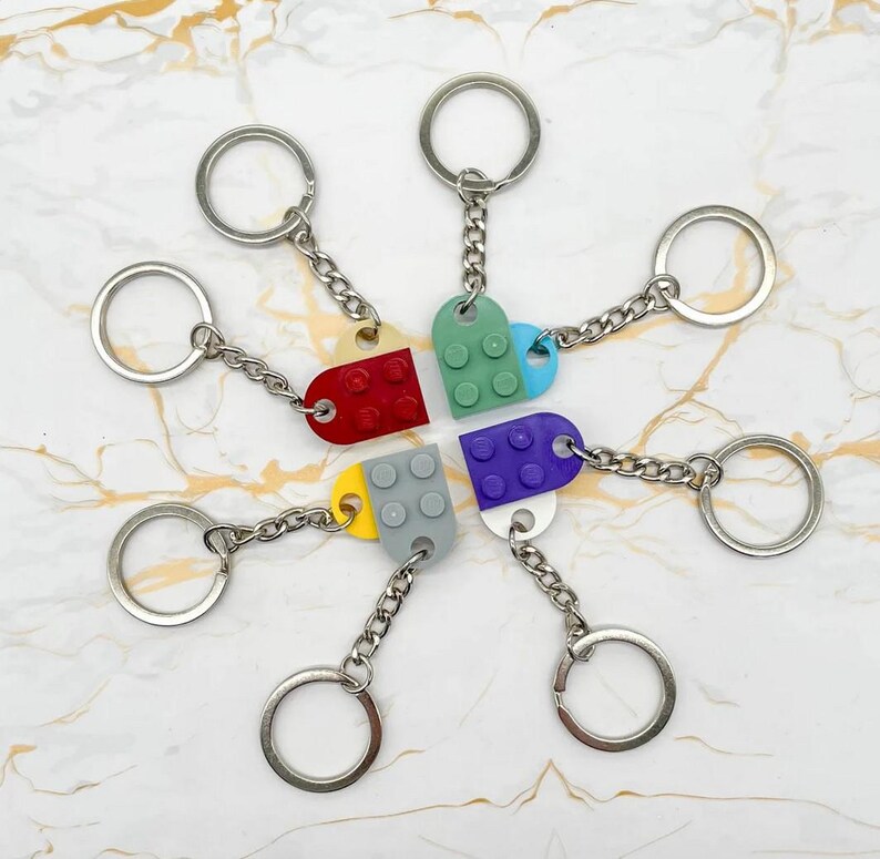 Pu&ograve; includere: Una collezione di portachiavi colorati, ognuno con un piccolo ciondolo a forma di mattone in rosso, verde, blu, viola, giallo e grigio. I ciondoli sono collegati a una catena d'argento e a un portachiavi rotondo. I portachiavi sono disposti a cerchio.