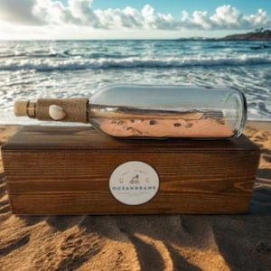 May include: A clear glass bottle with a cork stopper, twine, and a seashell, resting on a wooden box. The bottle contains sand and is labeled "OCEANGRAMS". The scene is set on a sandy beach with the ocean in the background.