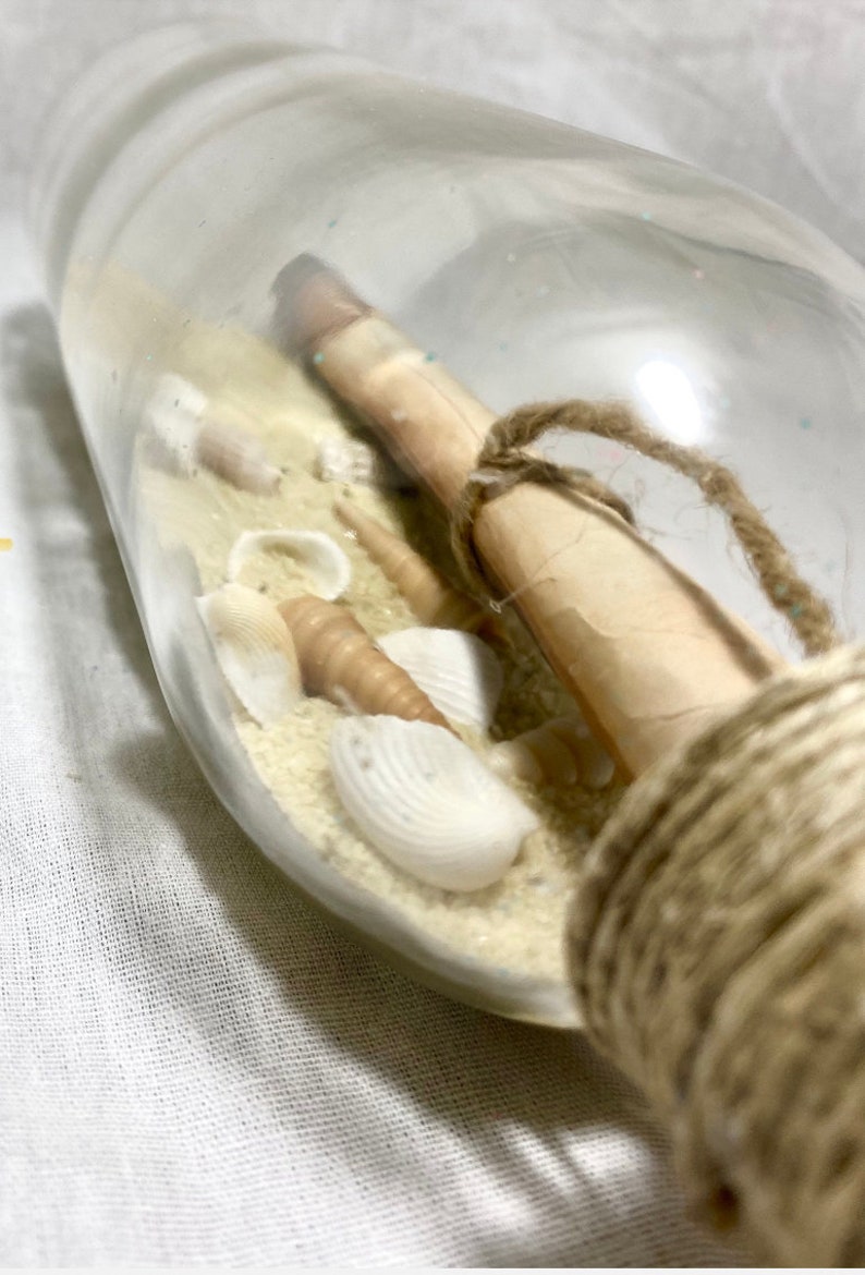 May include: A clear glass bottle containing sand, seashells, and a rolled-up scroll tied with twine. The bottle is wrapped with twine around the neck. The seashells are white and tan. The sand is light beige.