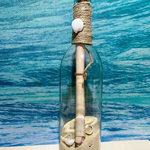 May include: A clear glass bottle with a cork stopper, twine, and a seashell. The bottle contains sand, a rolled-up scroll, and small seashells. The bottle is decorated with twine and sits against a blue and white background.