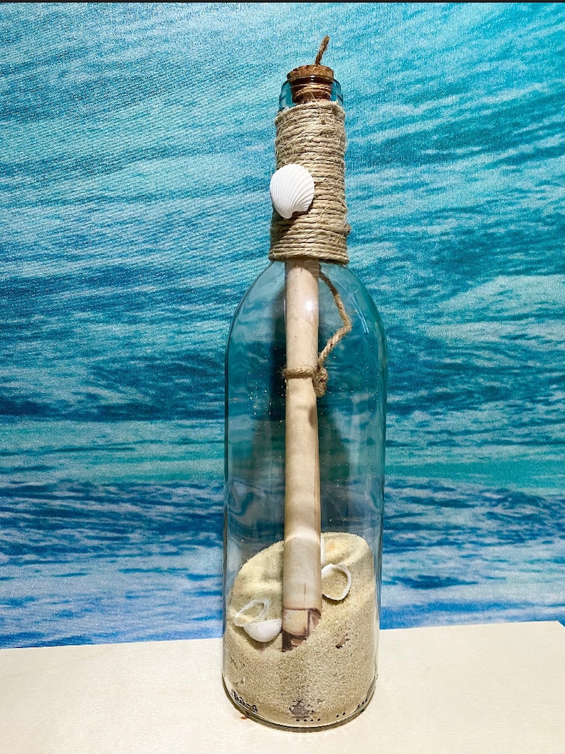 May include: A clear glass bottle with a cork stopper, twine, and a seashell. The bottle contains sand, a rolled-up scroll, and small seashells. The bottle is decorated with twine and sits against a blue and white background.