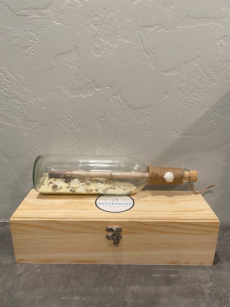 May include: A clear glass bottle with a cork stopper, twine, and a seashell detail, containing a rolled-up message and sand. The bottle rests on a wooden box with a metal clasp. The box has a sticker that says "OCEANGRAMS".