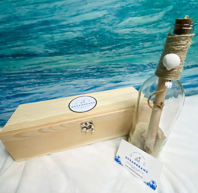 May include: A clear glass bottle with a cork stopper, twine, and a seashell, containing a rolled-up message. Beside it is a light wooden box with a silver clasp and a business card. The text on the business card and box reads "OCEANGRAMS".
