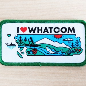 May include: A green and white embroidered patch with the text "I ♥ WHATCOM" and a stylized illustration of a landscape with mountains, a river, a fish, a boat, and trees.
