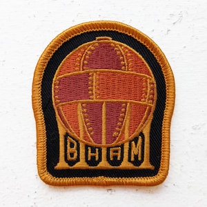 May include: A gold embroidered patch with a black background featuring a stylized image of a hot air balloon with the word "BHAM" underneath.