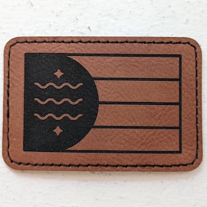 May include: A rectangular, brown leather patch with a black stitched border. The patch features a black design of a half-circle, wavy lines, and stars. The design is on a brown background with horizontal lines.
