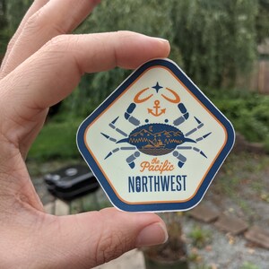 May include: A beige and blue diamond-shaped sticker with a stylized crab illustration. The crab is holding a small boat with mountains and a crescent moon in the background. The text "the Pacific NORTHWEST" is printed in blue and orange.