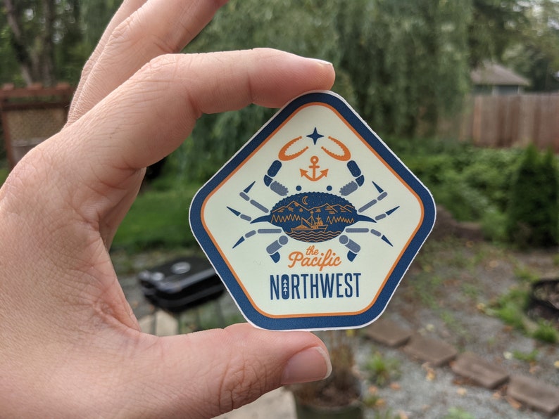 May include: A beige and blue diamond-shaped sticker with a stylized crab illustration. The crab is holding a small boat with mountains and a crescent moon in the background. The text "the Pacific NORTHWEST" is printed in blue and orange.