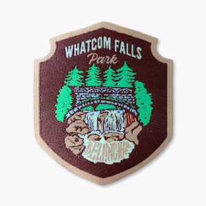 May include: A maroon and tan patch featuring a scenic illustration of Whatcom Falls Park in Bellingham. The design includes a waterfall, bridge, and evergreen trees, with the park name prominently displayed.