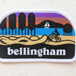 May include: An embroidered patch featuring a sunset scene, a sailboat, a person riding a bicycle, and the text "bellingham".