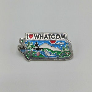 Könnte beinhalten: Eine silberne Anstecknadel mit einem blauen und grünen Landschaftsdesign. Die Nadel zeigt "I ♥ WHATCOM" mit einem roten Herzen. Die Landschaft zeigt einen Berg, eine Sonne, ein Boot, einen Fisch und eine Himbeere.