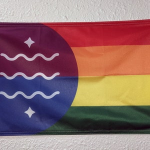 Puede incluir: Una bandera con un círculo azul que contiene tres líneas onduladas blancas y dos estrellas blancas. El círculo está sobre un fondo morado. El resto de la bandera es un arcoíris con rayas rojas, naranjas, amarillas, verdes y azules.