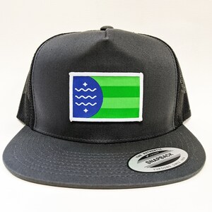 May include: A dark gray snapback hat with a mesh back and a flat brim. The hat features a rectangular patch with a blue and green design, including wavy lines and stars. The hat has a white border around the patch.