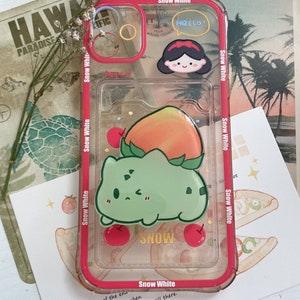 May include: A clear phone case with a cartoon peach and a green creature with the word "SNOW" printed on it. The case has a red border and the words "Snow White" printed on the sides. The case also has a sticker of a cartoon girl with the word "HELLO!" in a speech bubble.