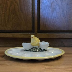 May include: A ceramic egg plate with a yellow rim and a decorative yellow bird figurine. The bird sits atop a small dish with a bed of dark green pebbles, and two small egg cup holders.