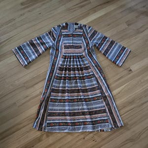 May include: A long-sleeved dress with a striped pattern in shades of blue, brown, and orange. The dress features a pleated bodice and a vertical stripe design down the front. The sleeves are wide and the dress is made of a lightweight fabric.