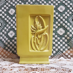 May include: A yellow ceramic vase with a raised design of a bird of paradise flower. The vase has a square shape with a rectangular opening at the top.