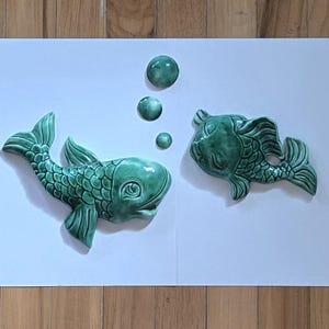 May include: Two teal ceramic fish sculptures with bubble accents on a white background. The fish are facing each other, with detailed scales and fins. The artwork is displayed on a wooden surface.