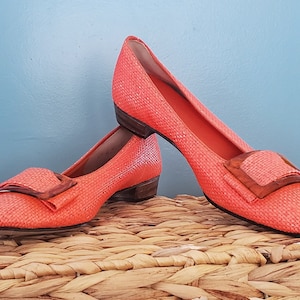 May include: A pair of coral-colored woven shoes with a brown buckle. The shoes have a low heel and are sitting on a woven basket.