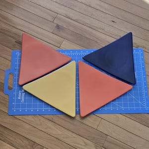 May include: Four colorful triangular shapes in orange, yellow, and navy blue are arranged on a blue cutting mat. The mat has grid lines and measurement markings. The shapes are on a wooden surface.