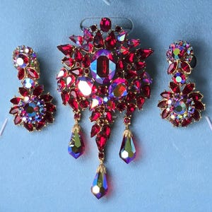 May include: A vintage jewelry set featuring a large brooch and matching earrings. The set showcases ruby-red and iridescent crystals set in gold-tone metal. The brooch has dangling teardrop-shaped pendants. The earrings have a floral design.