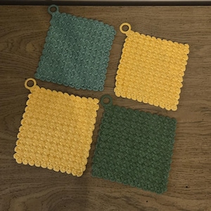 May include: Four square crochet potholders in shades of teal, yellow, and green, each with a loop for hanging. The potholders are arranged on a wooden surface, showcasing their textured, handmade appearance.
