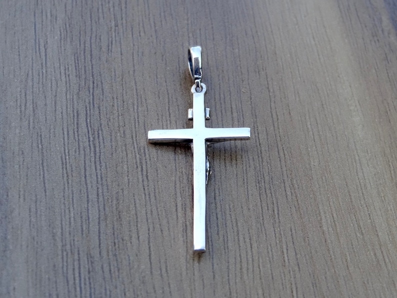 Puede incluir: Pendiente de plata en forma de cruz con un dise&ntilde;o detallado. La cruz est&aacute; sobre un fondo de madera.