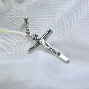 Puede incluir: Un colgante de plata en forma de cruz con una figura detallada de Jesucristo. El colgante est&aacute; sobre un fondo de tela blanca.